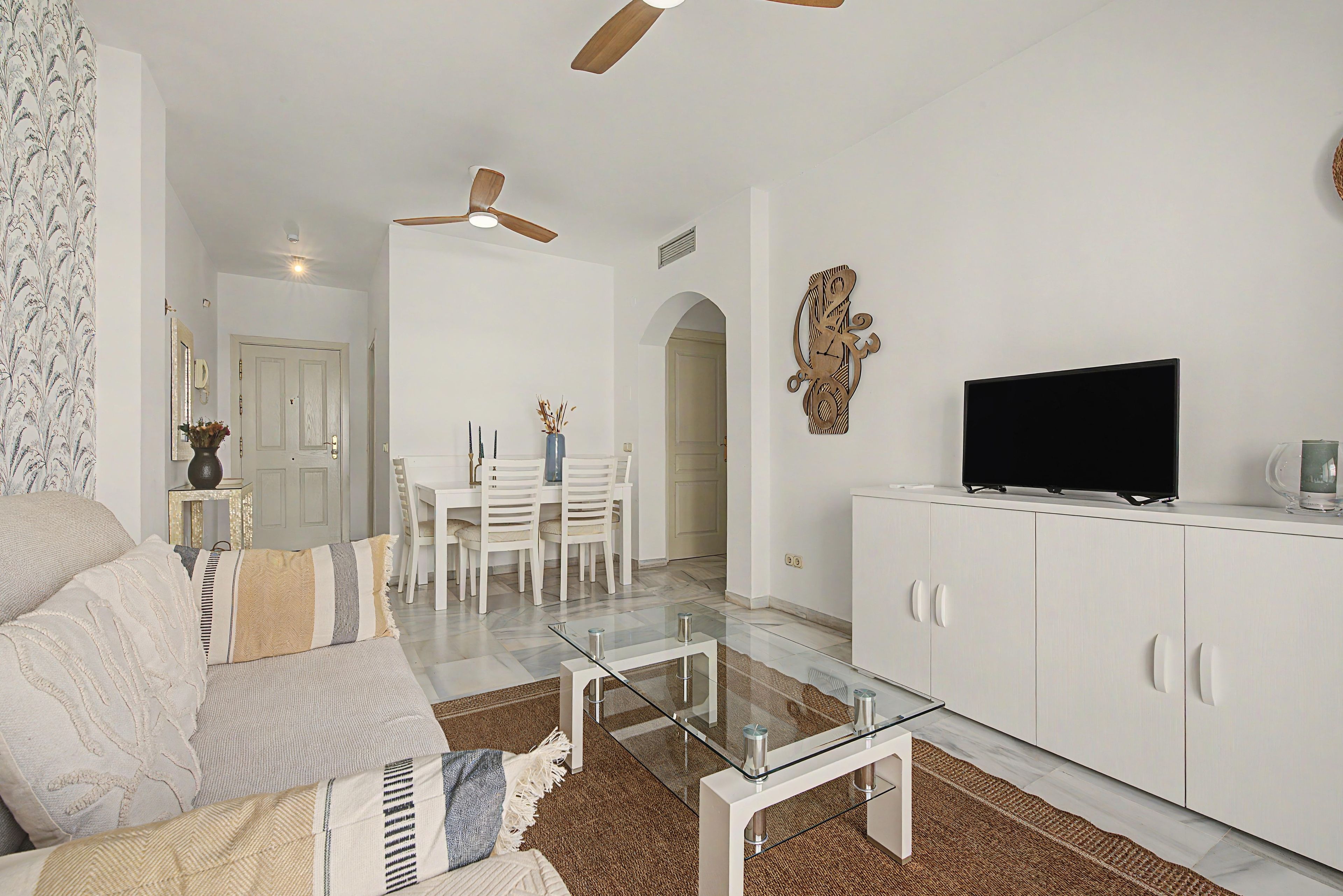 A modern living room with a glass coffee table, white furniture, a ceiling fan, and a flat-screen TV on a cabinet.