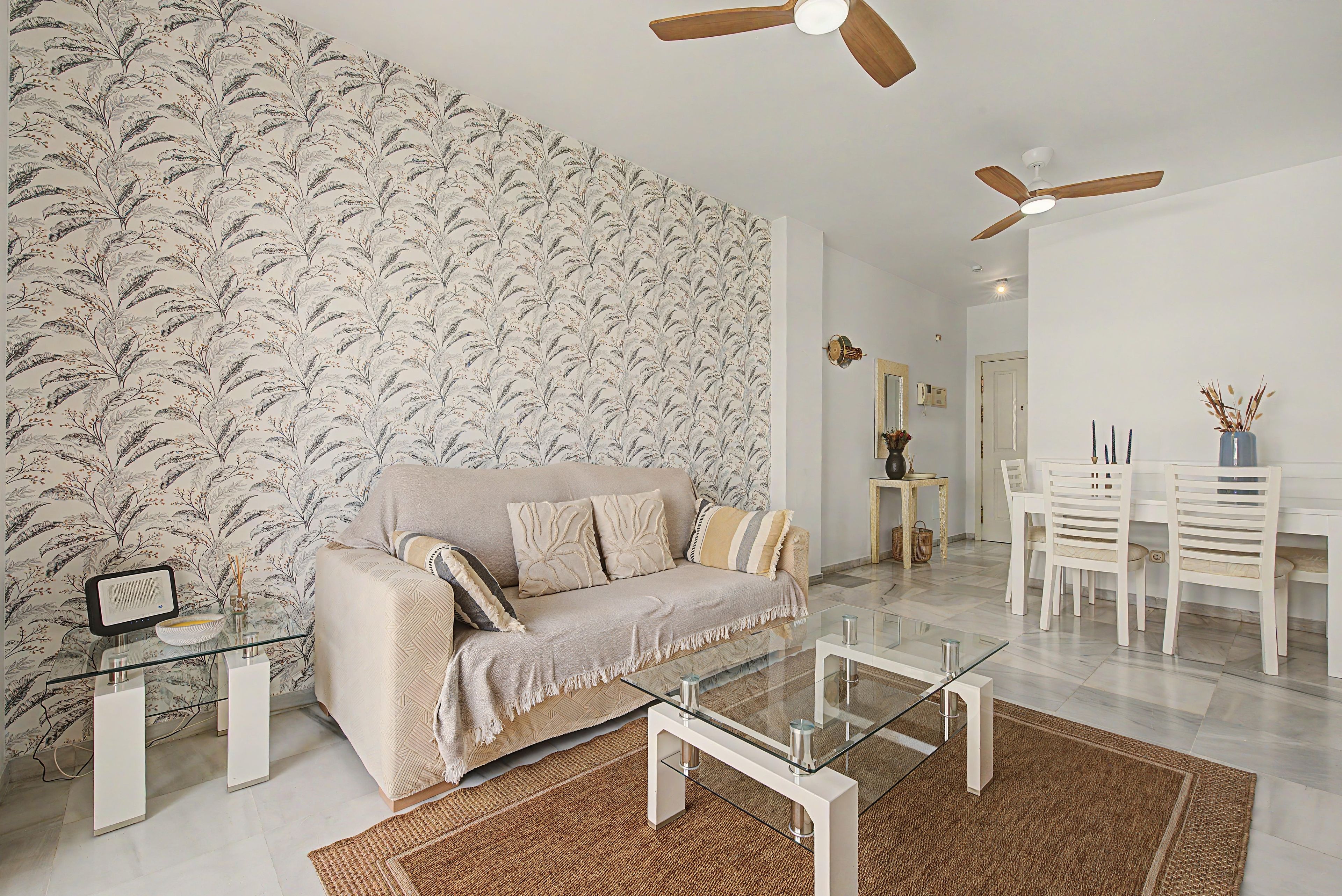 Uma sala de estar moderna com papel de parede estampado, um sofá bege, uma mesa de centro de vidro e um ventilador de teto.