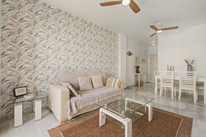 A modern living room with a patterned wallpaper, a beige sofa, a glass coffee table, and a ceiling fan.