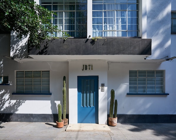 Un edificio con una porta blu con la scritta "JB71", due cactus in vaso e un balcone con piante.
