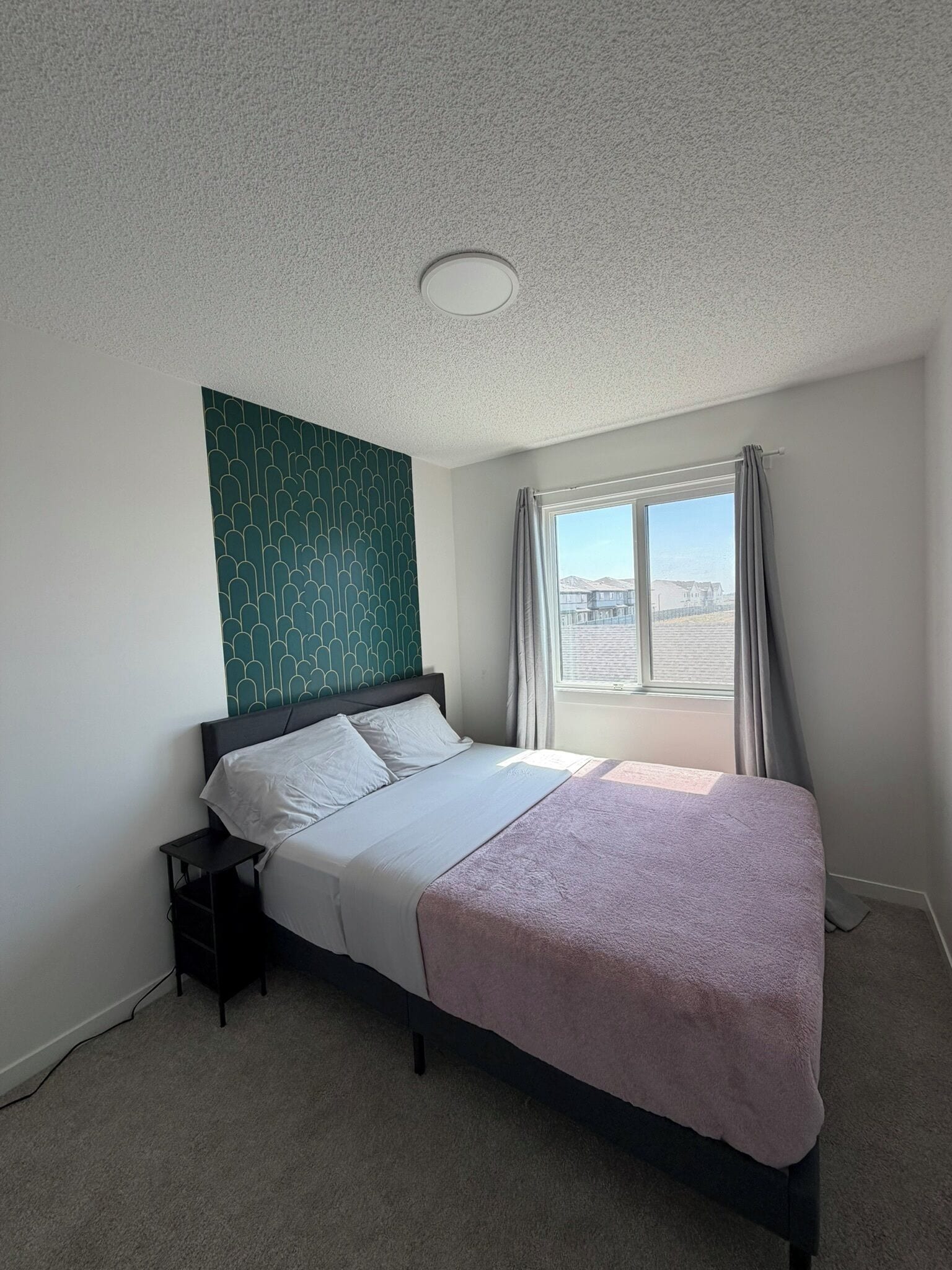 A bedroom with a bed, a nightstand, a window with curtains, and a patterned wall.