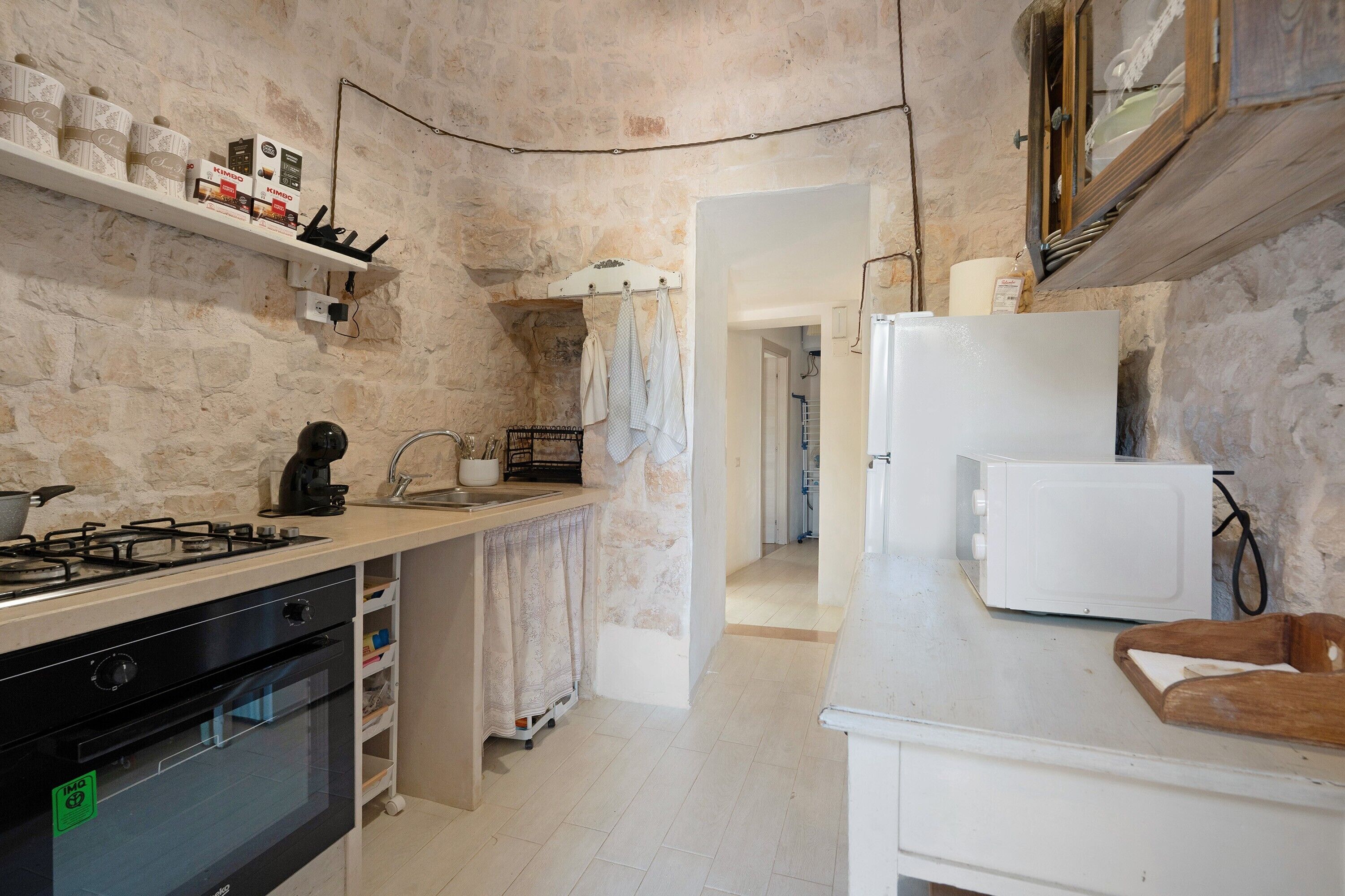 A kitchen with a stove, oven, and white refrigerator. There is a shelf with boxes and a hanging rack with cloths.