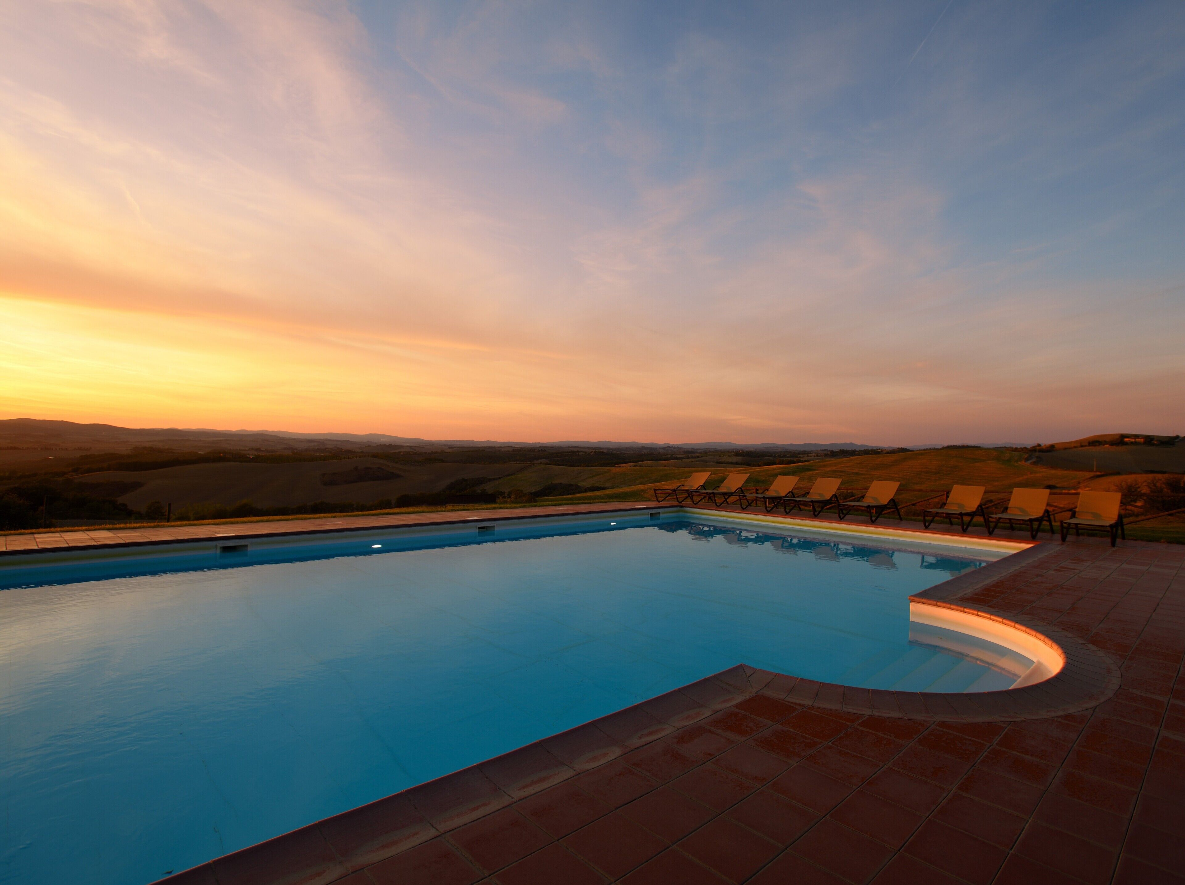 Una piscina con borde curvo, rodeada de sillas de descanso y vistas a colinas ondulantes.
