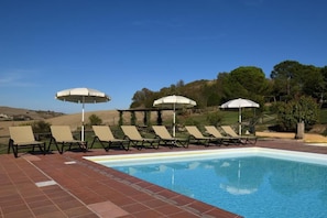 Una piscina amb gandules i ombrel·les, envoltada d'una zona ajardinada amb arbres i turons.