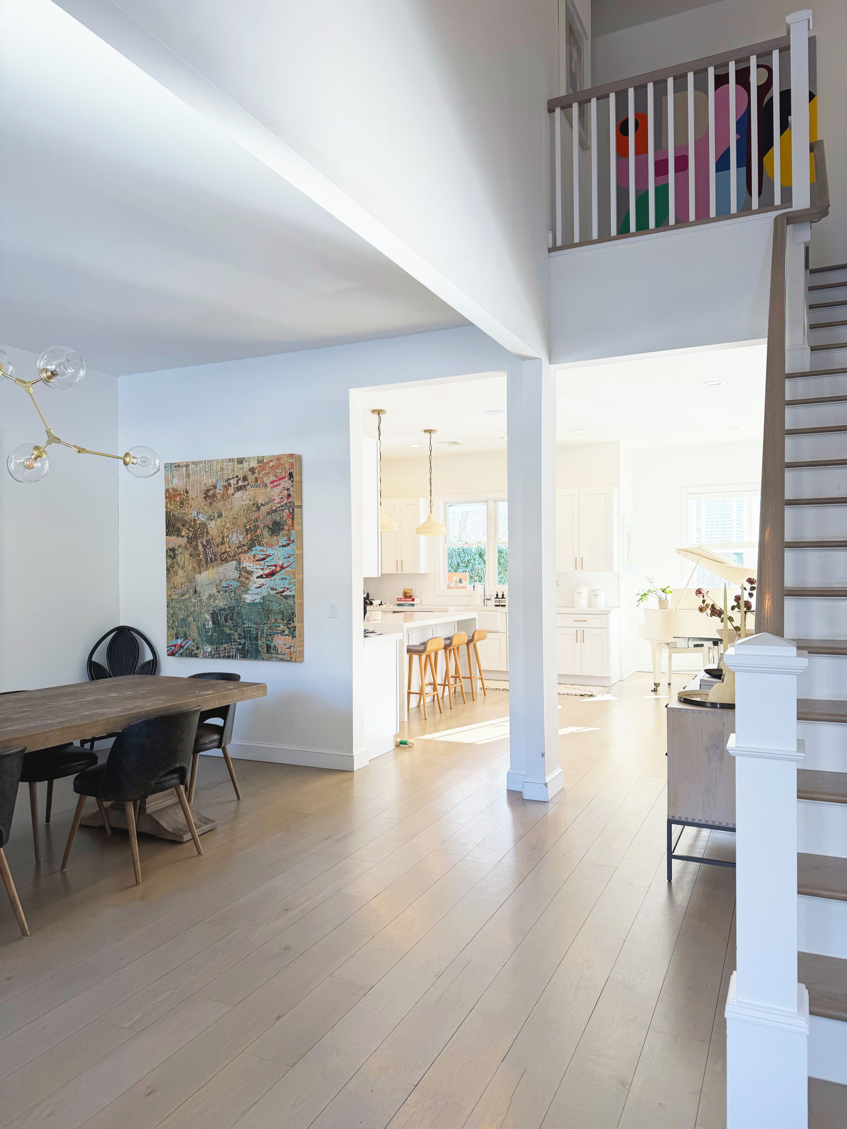 Un vaste espace de vie comprenant une table à manger en bois, des chaises noires, un grand tableau abstrait et un escalier avec une rampe blanche.