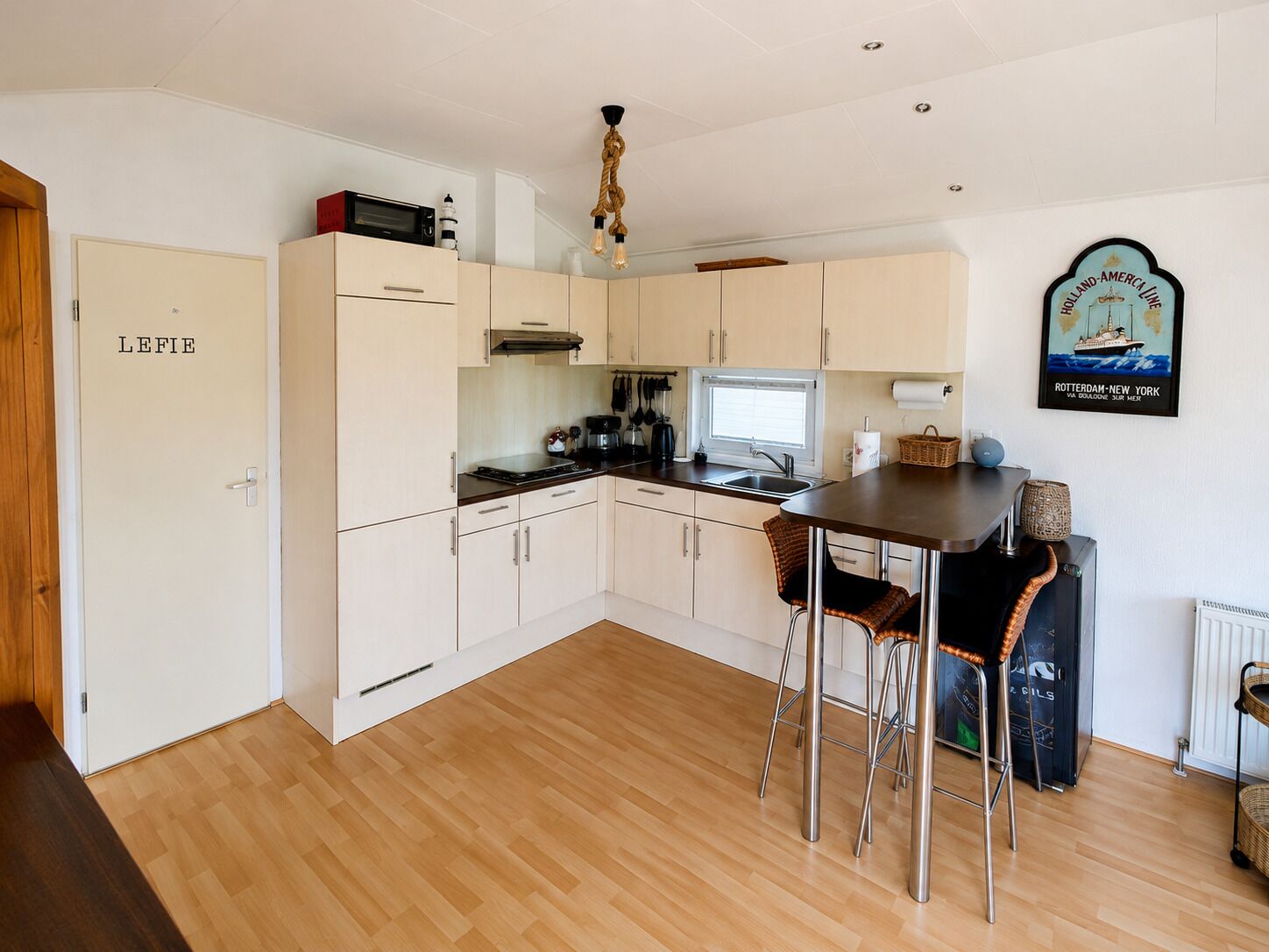 A compact kitchen with white cabinets, a wooden floor, a dining area with two chairs, and a wall-mounted light fixture.