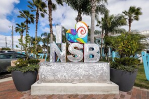 Ein Schild mit den Buchstaben "NSB" und einem farbenfrohen Herzdesign, umgeben von Palmen und Topfpflanzen.