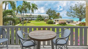 A balcony with a round wooden table and wicker chairs, overlooking a well-maintained lawn, a pool, and a building with a view of the ocean. - Awesome Beach Condo on Perfect Seven Mile Beach! (Seven Mile Beach)