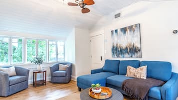 Un salon avec un canapé bleu, deux fauteuils, une table basse avec un plateau, un ventilateur de plafond et un tableau accroché au mur.