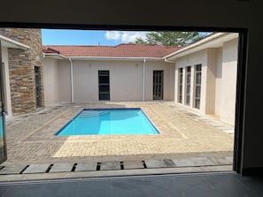 A swimming pool with a tiled deck, surrounded by walls with windows and a stone pillar.