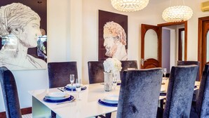 Uma sala de jantar com uma mesa branca posta para seis pessoas, rodeada por cadeiras estofadas escuras. Há dois grandes quadros pendurados na parede, um vaso com flores brancas e um candelabro.