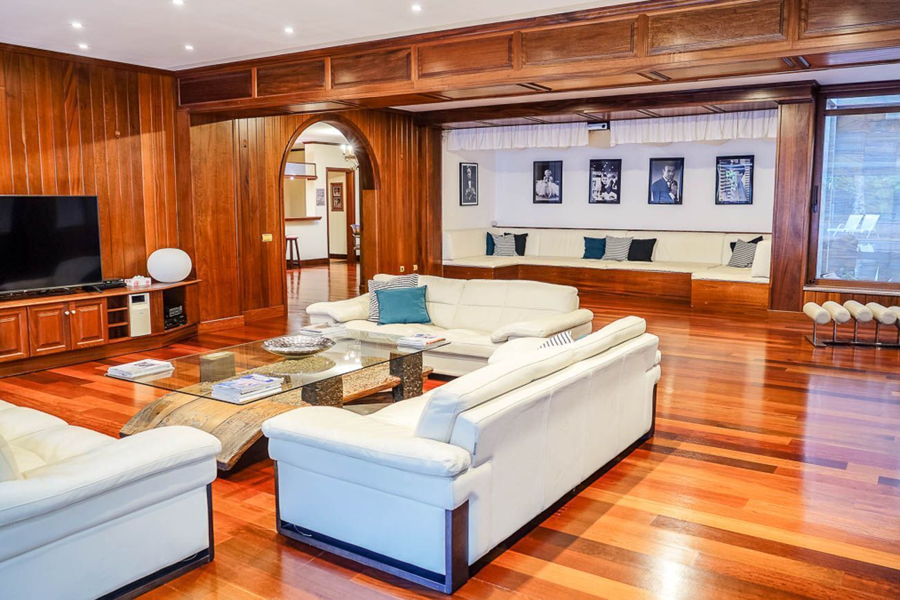 A spacious living room with a large flat-screen TV, a glass coffee table, and a white sofa set.