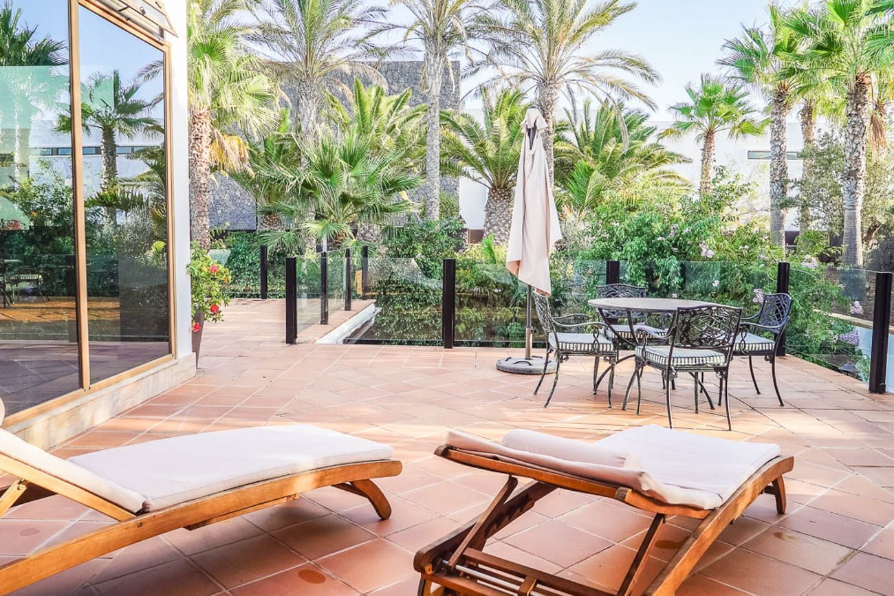 A sunlit outdoor area with lounge chairs, a table, and chairs, surrounded by palm trees and a glass wall.