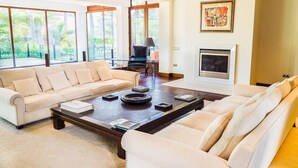 Uma sala de estar ampla com um sofá bege, uma mesa de centro com livros e objetos decorativos, uma lareira e grandes janelas com vista para o verde.