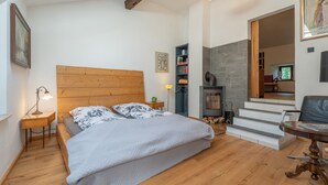 A bedroom with a wooden headboard, a bed with floral bedding, a nightstand with a lamp, a bookshelf, a fireplace, and a chair. - Holiday Apartment in Kipfenberg – 1 Bedroom / Sleeping Gallery / 2 Bathrooms (Kipfenberg)