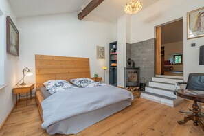 Ett sovrum med en sänggavel i trä, en säng med blommigt sänglinne, ett nattduksbord med en lampa, en bokhylla, en öppen spis och en stol.