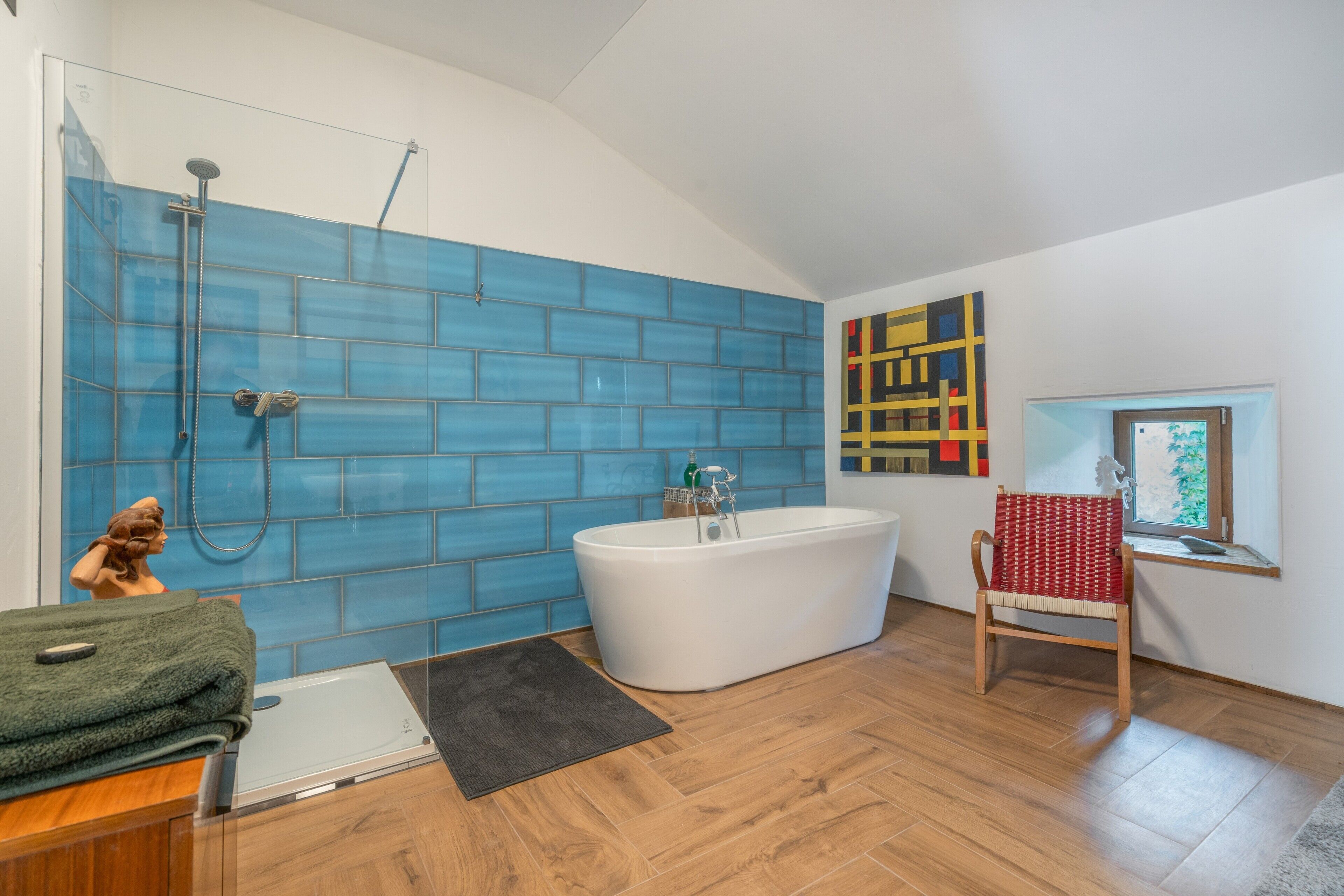 A modern bathroom with a blue-tiled shower wall, a freestanding bathtub, a wooden floor, and a red and white patterned chair.