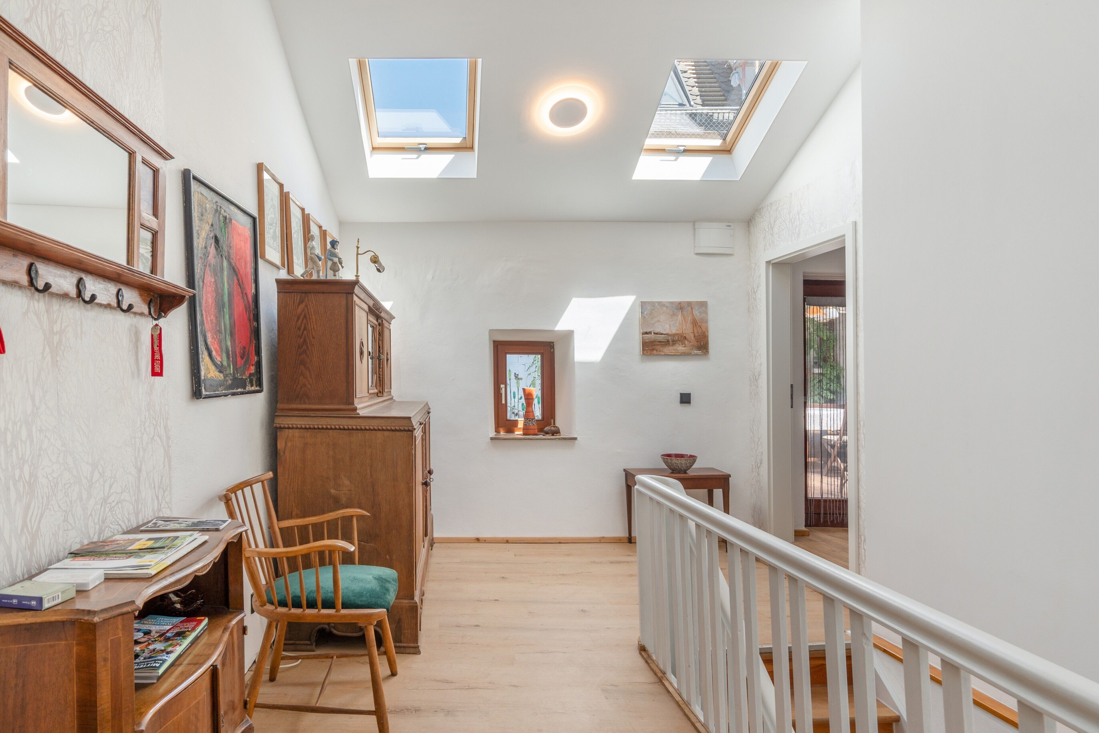 A hallway with wooden flooring, a wooden chair, a desk with books, and a cabinet. There are skylights and a window with a view.