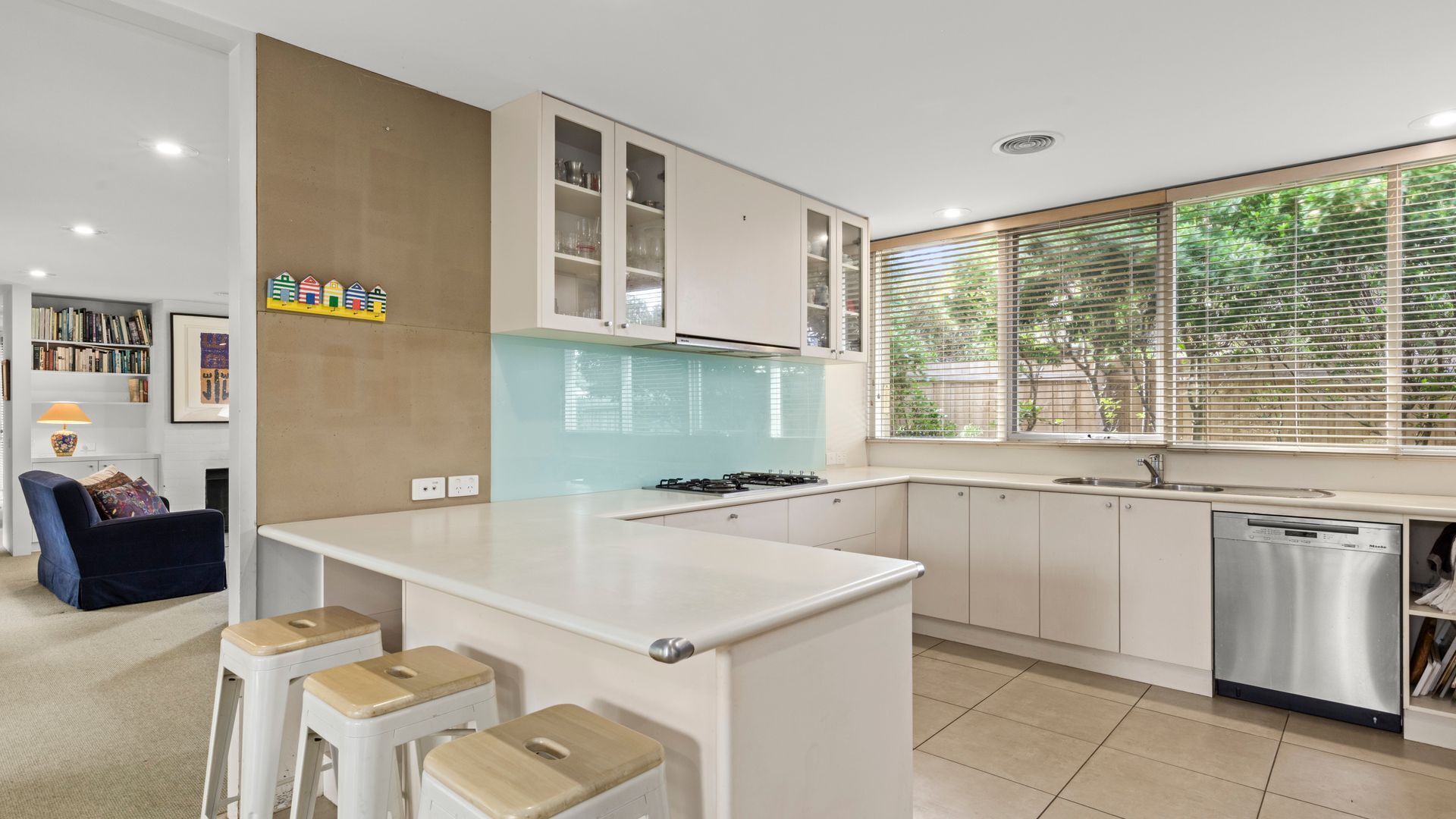 A modern kitchen with a central island, bar stools, a dishwasher, and a window with blinds.