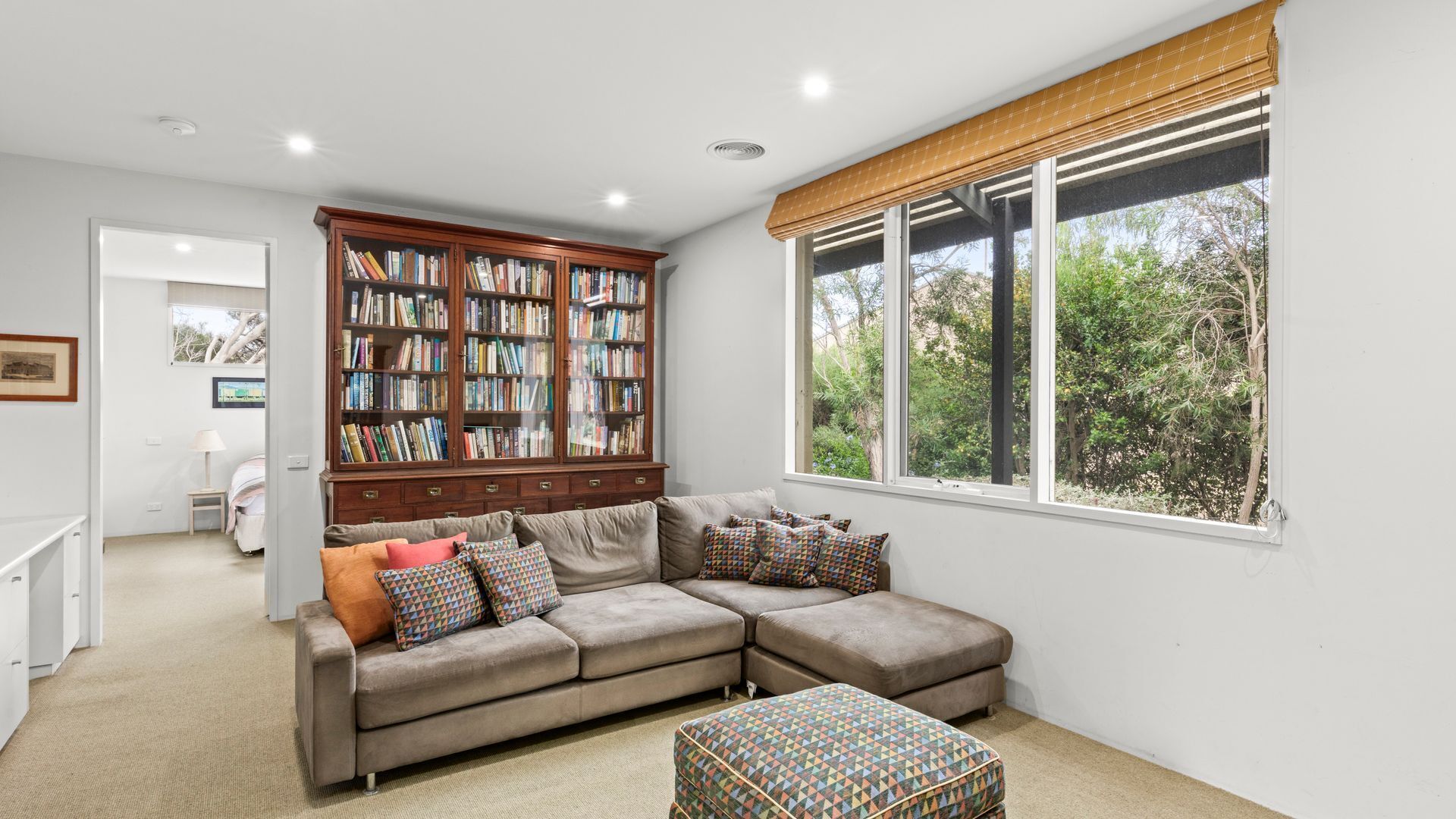 A living room with a sectional sofa, a large bookshelf, and a window with a view of trees.