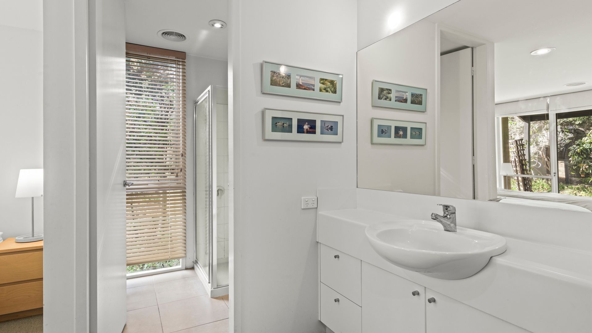 Een moderne badkamer met een grote spiegel, een witte wastafel en een raam met zonwering.