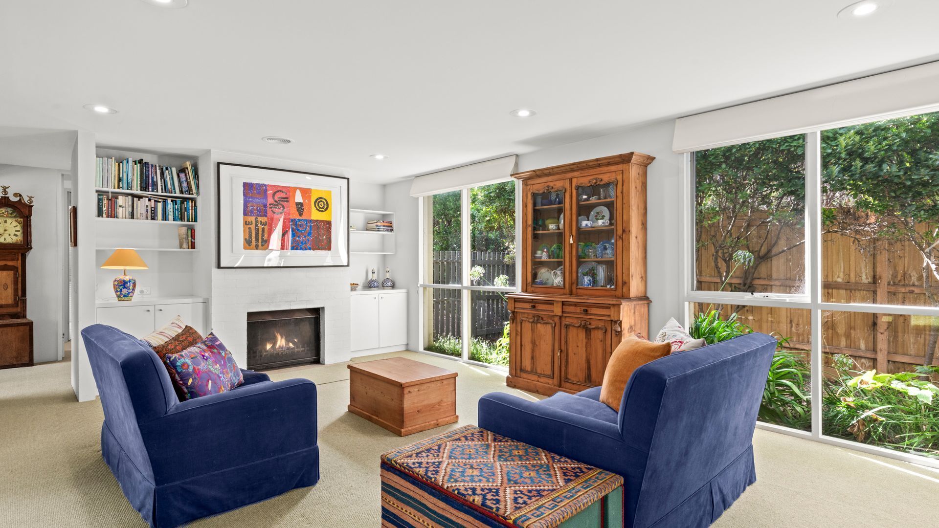 A living room with two blue sofas, a fireplace, a bookshelf, and a large window with a view of greenery.