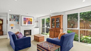 A living room with two blue sofas, a fireplace, a bookshelf, and a large window with a view of greenery. - The Quiet Nook - Point Lonsdale (Point Lonsdale)