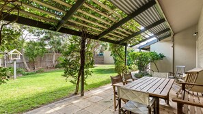Ein überdachter Patio-Bereich mit Holztisch und Stühlen, eine Pergola mit Kletterpflanzen und Blick auf einen Garten mit Zaun und Bäumen.