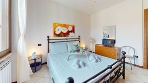 Une chambre bien rangée, avec un lit, une table de chevet, une petite table avec une lampe, un ventilateur, un téléviseur et un tableau accroché au mur.