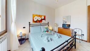 Ein ordentlich eingerichtetes Schlafzimmer mit einem Bett, einem Nachttisch, einem kleinen Tisch mit einer Lampe, einem Ventilator, einem Fernseher und einem Bild an der Wand.