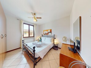 Een hotelkamer met een bed, nachtkastje, bureau, stoel, plafondventilator, raam, radiator, kunstwerk en een spiegel.