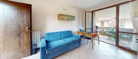 A living room with a blue sofa, a dining table with chairs, a sliding glass door to a balcony, and a wooden door.