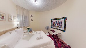 Une chambre d’hôtel avec un lit, une table de chevet avec une lampe, une petite table sur laquelle se trouve un plateau avec des articles pour le petit-déjeuner, un ventilateur et un tapis décoratif.