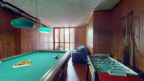 Ein Billardtisch mit Billardkugeln, ein Kicker und eine Couch in einem Raum mit Holzwänden und einer geneigten Decke.