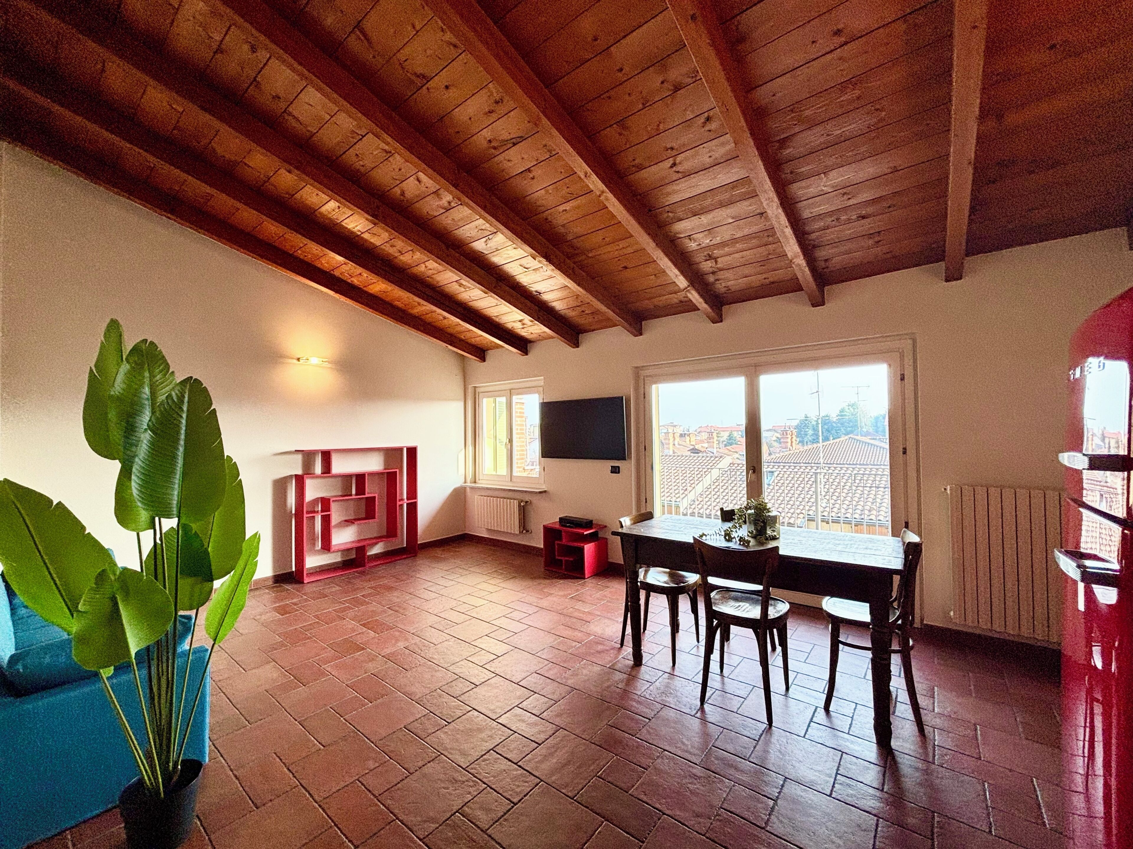 Une pièce spacieuse avec un plafond en bois, une table à manger avec des chaises, une étagère rouge, une plante en pot et une fenêtre donnant sur des immeubles.