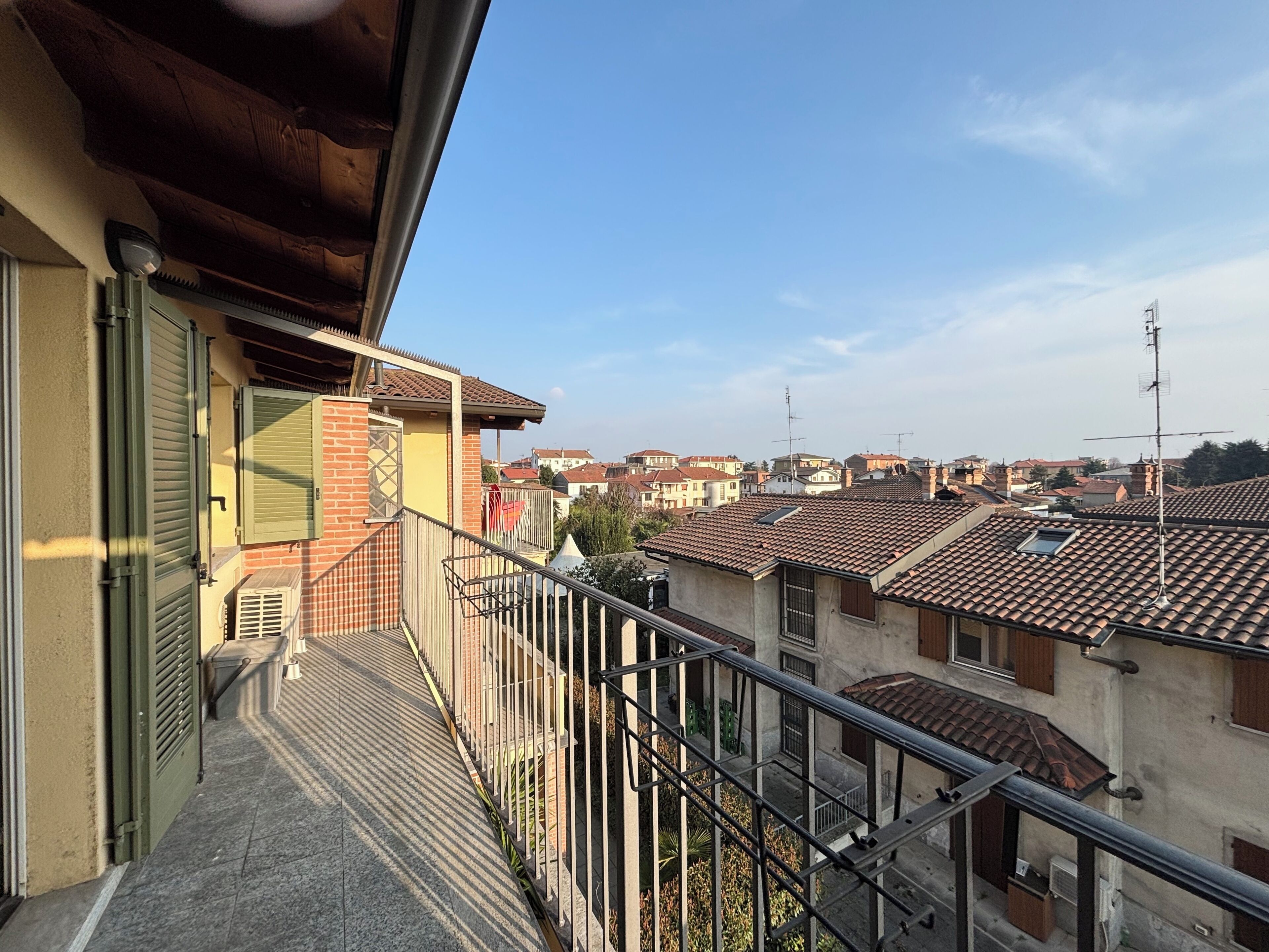 Un balcon donnant sur un quartier résidentiel, avec un toit en tuiles, une rambarde en métal et une porte équipée de volets.
