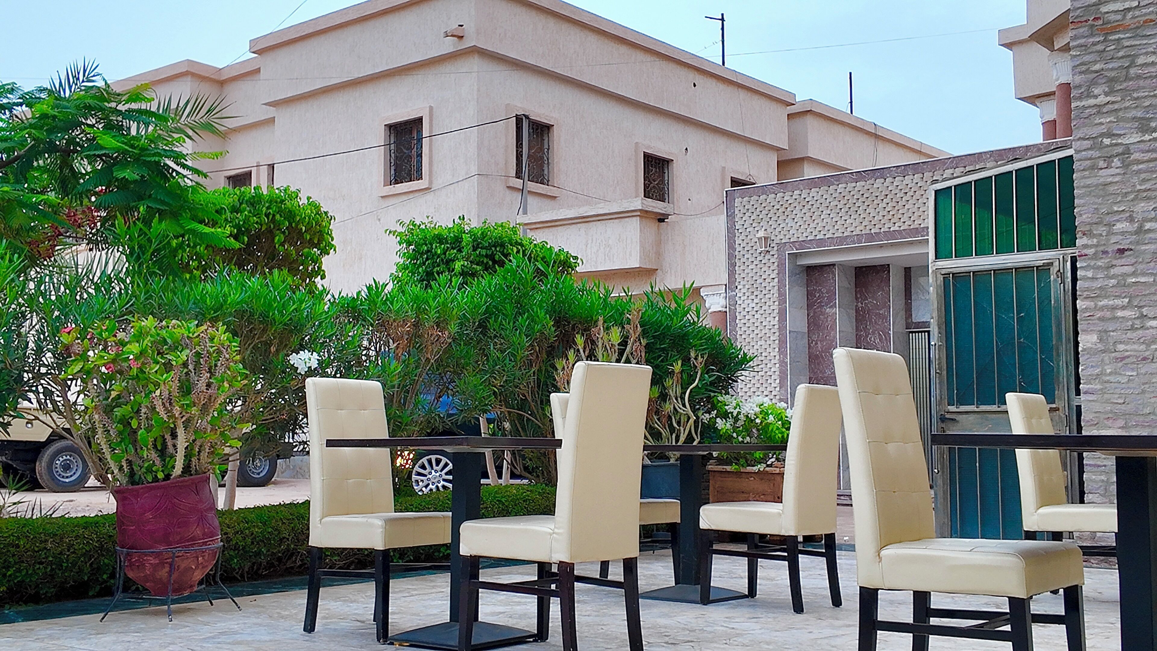 Outdoor seating with chairs and tables, surrounded by greenery and a building with a tiled facade.
