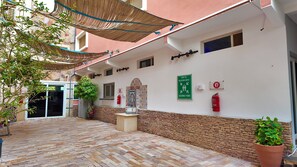 Un patio con suelo de ladrillo, techo de paja y pared de piedra. Hay plantas en macetas y un extintor de incendios fijado a la pared.