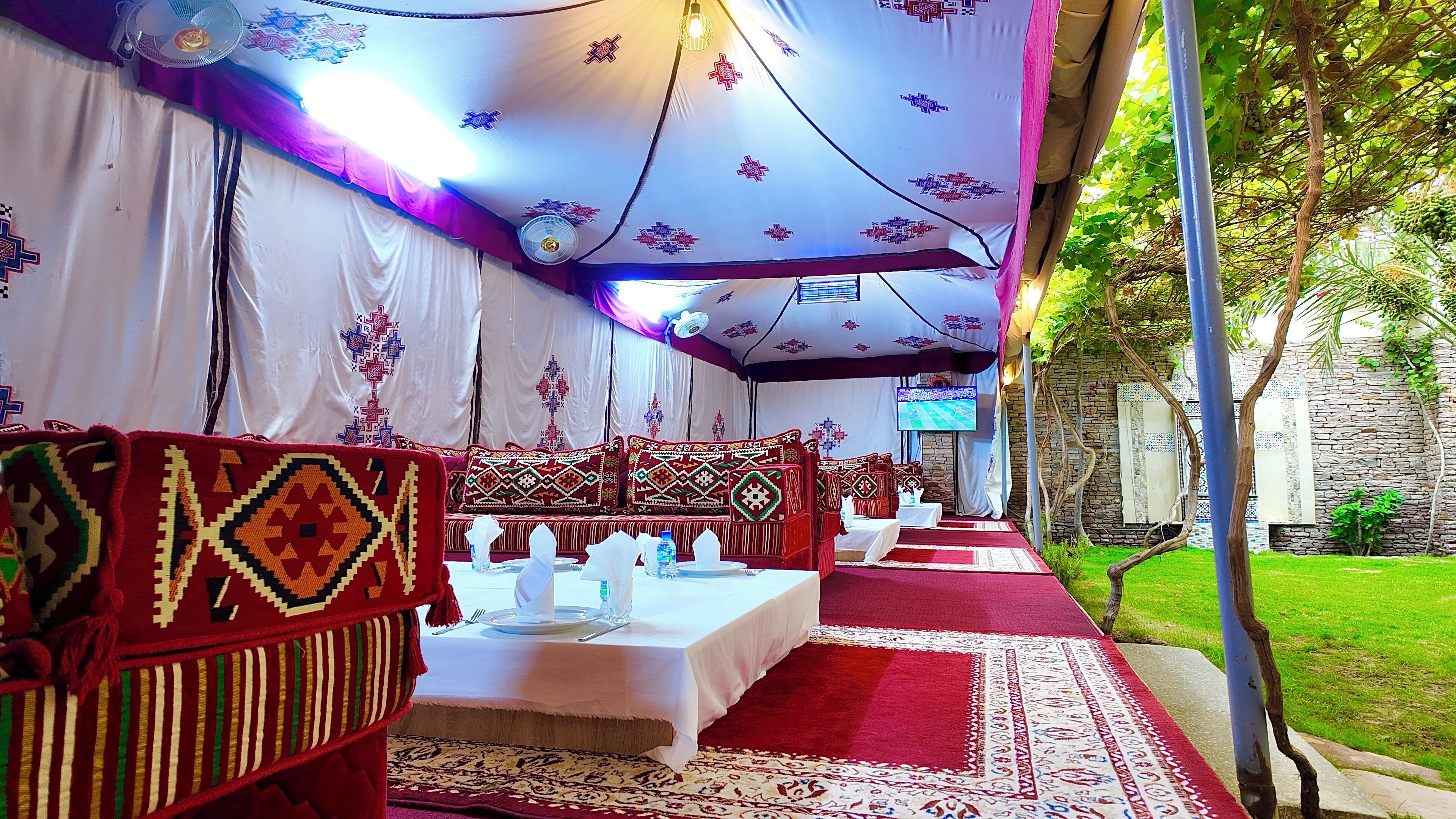 A tented dining area with tables set for a meal, chairs with patterned cushions, and a stone wall in the background.