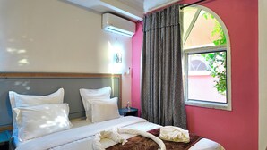 Habitación de hotel con cama, almohadas, mesita de noche, equipo de aire acondicionado, ventana con cortinas y vista a zonas verdes.