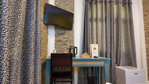 Una habitación con televisión de pantalla plana montada en una pared de ladrillo, una mesa con una silla, una cafetera y un refrigerador pequeño.