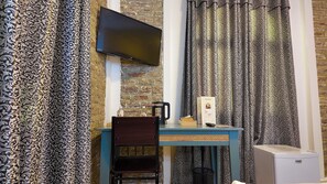 A room with a flat-screen TV mounted on a brick wall, a table with a chair, a coffee maker, and a small refrigerator.