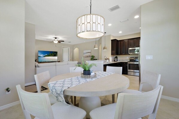 A dining area with a round table, chairs, a ceiling fan, and a kitchen in the background.
