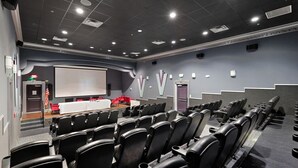 Een vergaderzaal met rijen zwarte stoelen, een wit projectiescherm en een podium met een rode tafel.