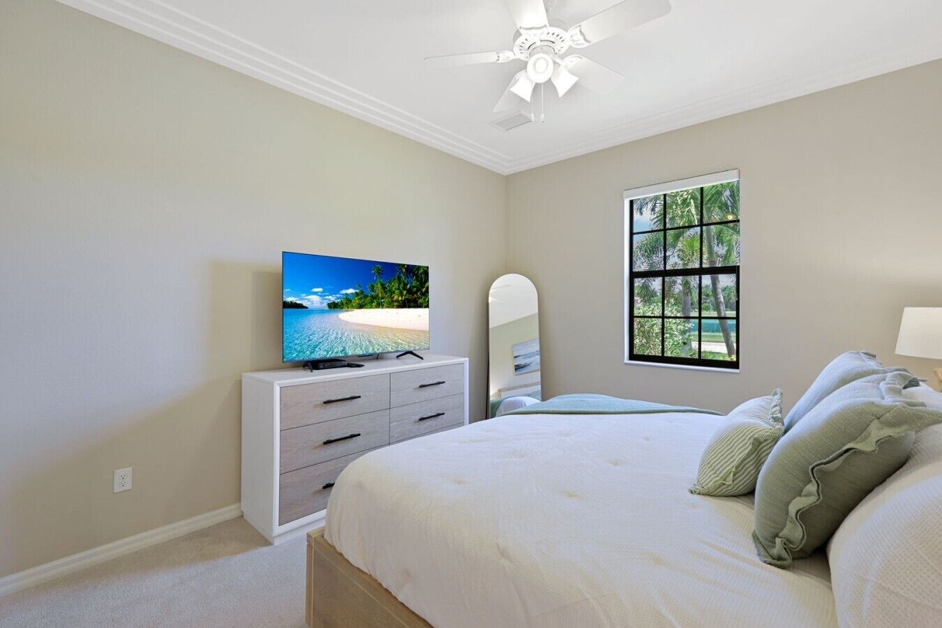 A bedroom with a bed, a television on a dresser, a window with a view of trees, and a ceiling fan.