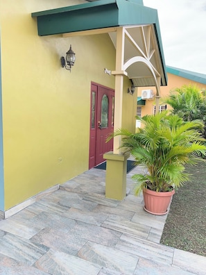 A building with a red door, a green awning, and a potted plant in the foreground. - Alluring Ambrosia (Arouca)