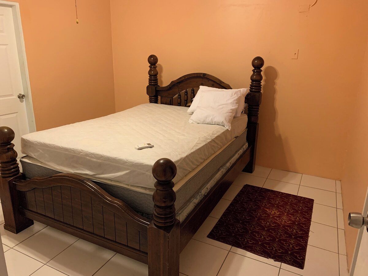 Un lit simple avec une tête de lit et un pied de lit en bois, un matelas blanc et un oreiller assorti. Un petit tapis est placé au pied du lit.