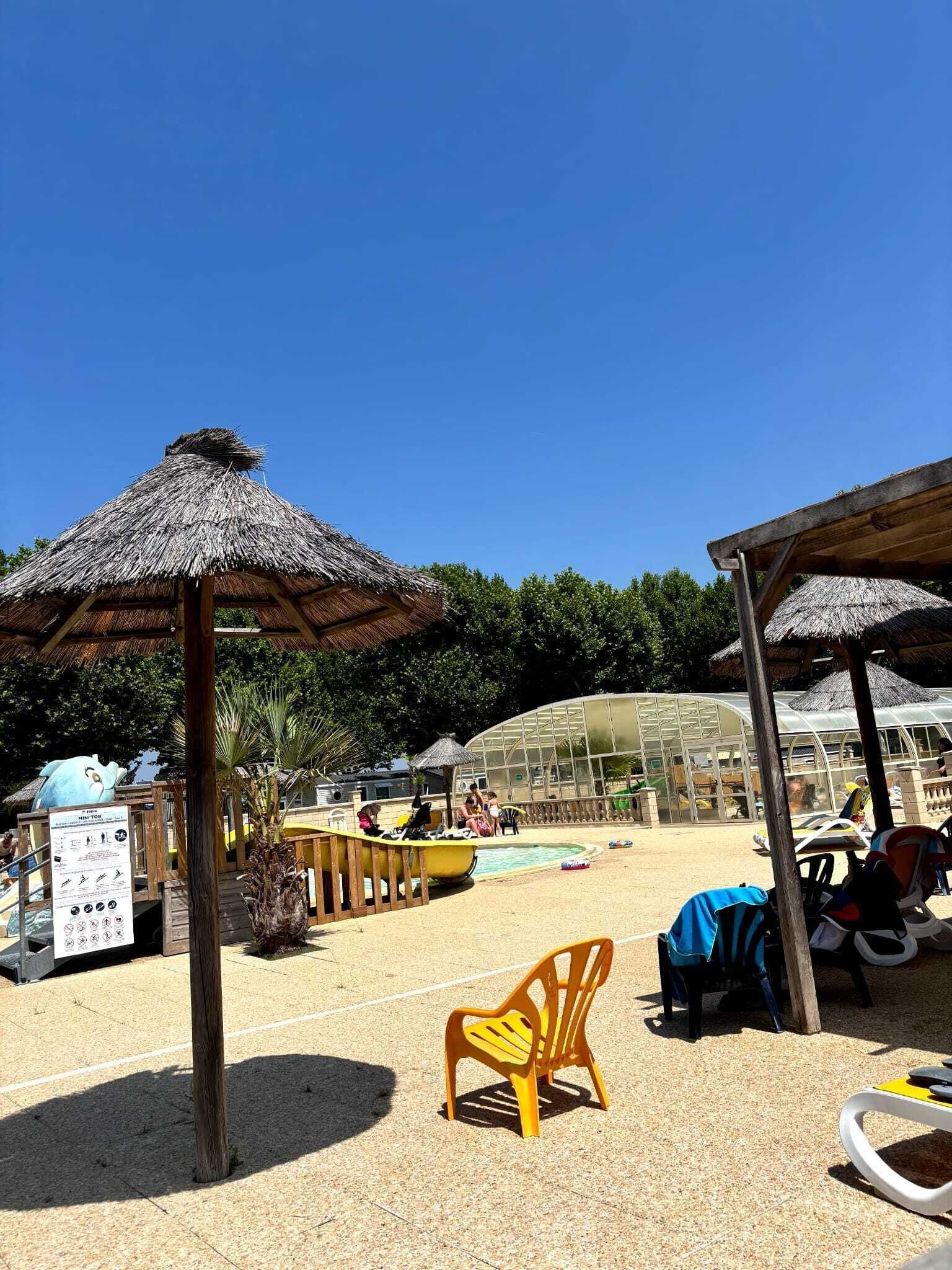 A beach area with thatched umbrellas, lounge chairs, a pool, and a greenhouse structure.