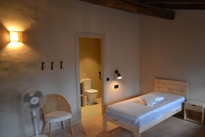 A bedroom with a bed, a chair, a fan, and a toilet visible through an open door. - Can Hortafa - Country House in Tortellà - Cortada de la Cellera (Tortellà)