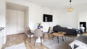 A modern living room with a dining area, a flat-screen TV mounted on the wall, and a bed with a dark-colored headboard. - Mall of Tripla apartments by Come in (Helsinki)
